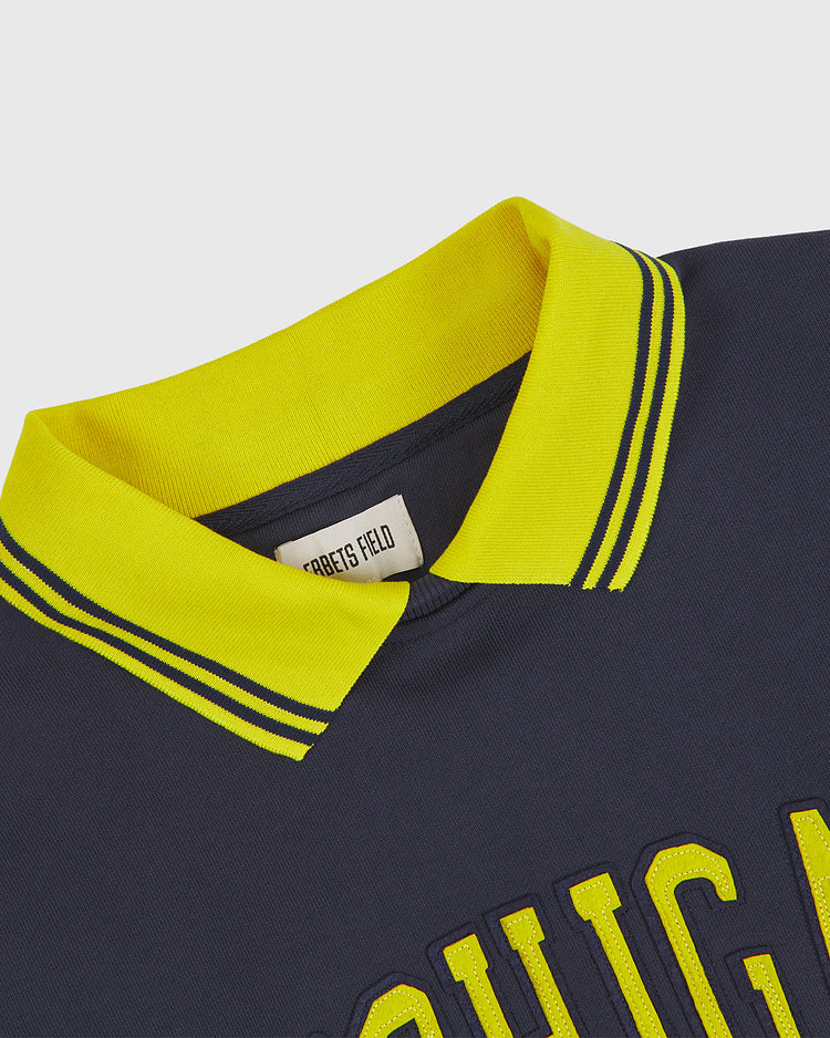 Close-up of the University of Michigan Collared Crewneck in navy blue with a bright yellow collar and striped trim. The visible label inside reads Ebbets Field. Yellow text is partially visible on the front of the shirt.