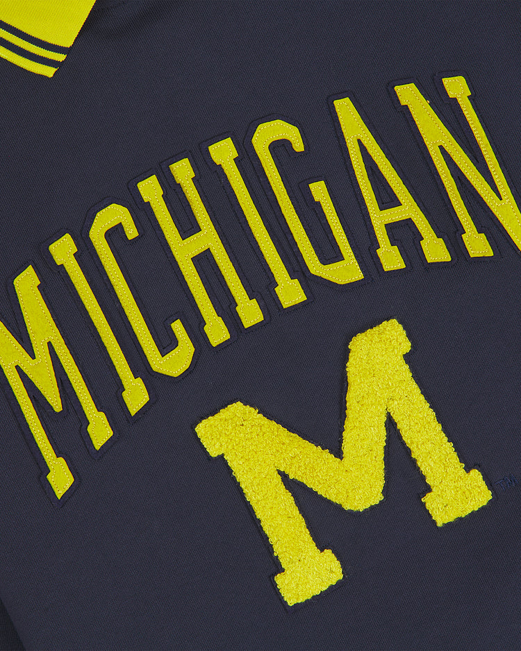 Close-up of the University of Michigan Collared Crewneck by Ebbets Field, featuring navy fabric with yellow, textured “Michigan” text and a large textured letter “M.”.