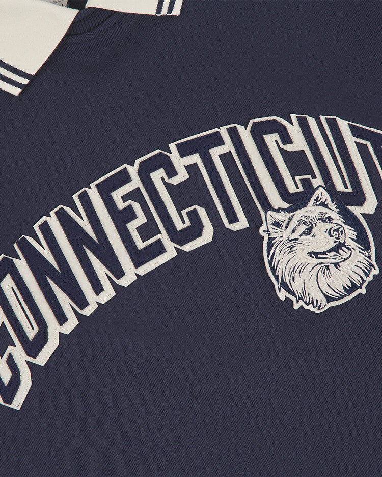 Ebbets Field University of Connecticut Collared Crewneck in navy blue, featuring large white “CONNECTICUT” text and a white husky dog head graphic below, with a visible white collar accented by navy stripes at the top.