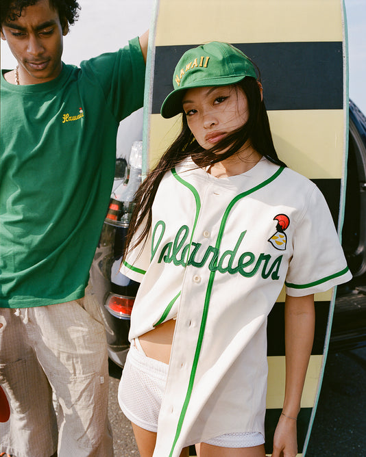 Two young adults stand in front of a car; the woman wears a white Islanders baseball jersey, white shorts, and a green Hawaii cap, leaning on a striped surfboard. The man wears a green Hawaii T-shirt and beige pants.