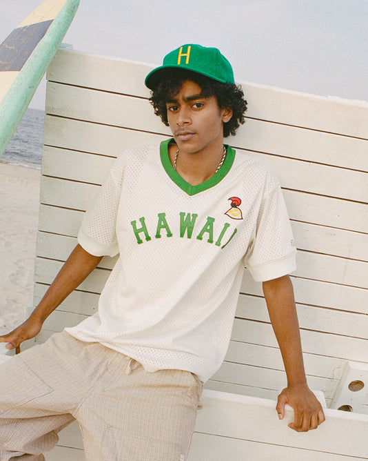 A young man in a green cap and white HAWAII jersey sits on a wooden bench at the beach, with a surfboard and ocean in the background.
