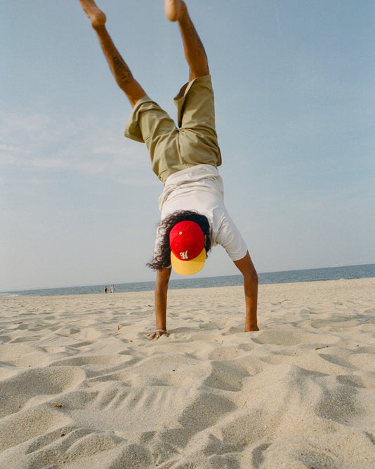 A person in a red and yellow cap, white shirt, and khaki shorts does a handstand on a sandy beach under a blue sky, with the ocean in the background.