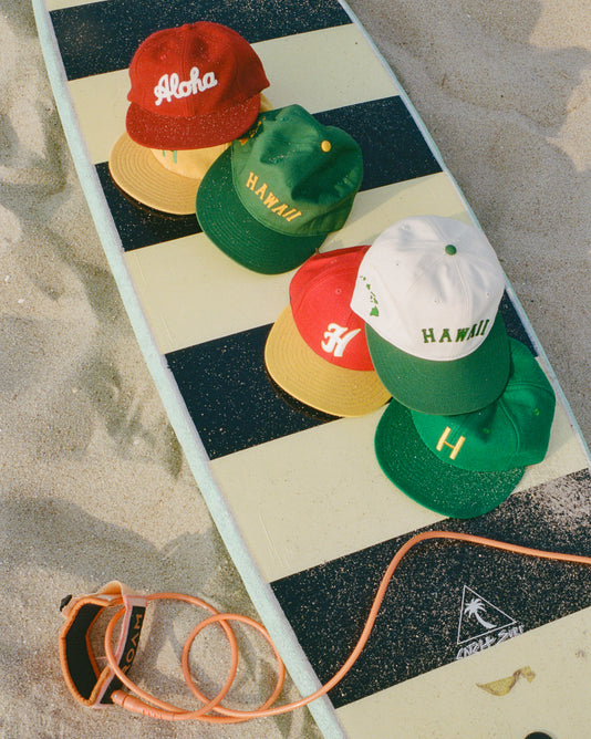 A striped surfboard on sand with five colorful caps on top, each featuring Hawaiian-themed text, and an orange surf leash attached to the board.