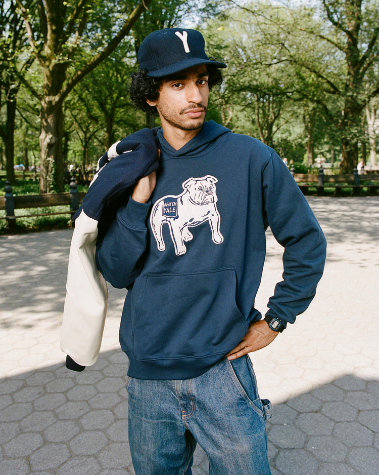 A man in a navy Yale University Heavyweight Cotton Terry Pullover Hoodie by Ebbets Field, blue jeans, and a dark Yale cap stands outdoors in a park with trees and a paved path, holding a jacket over his shoulder.