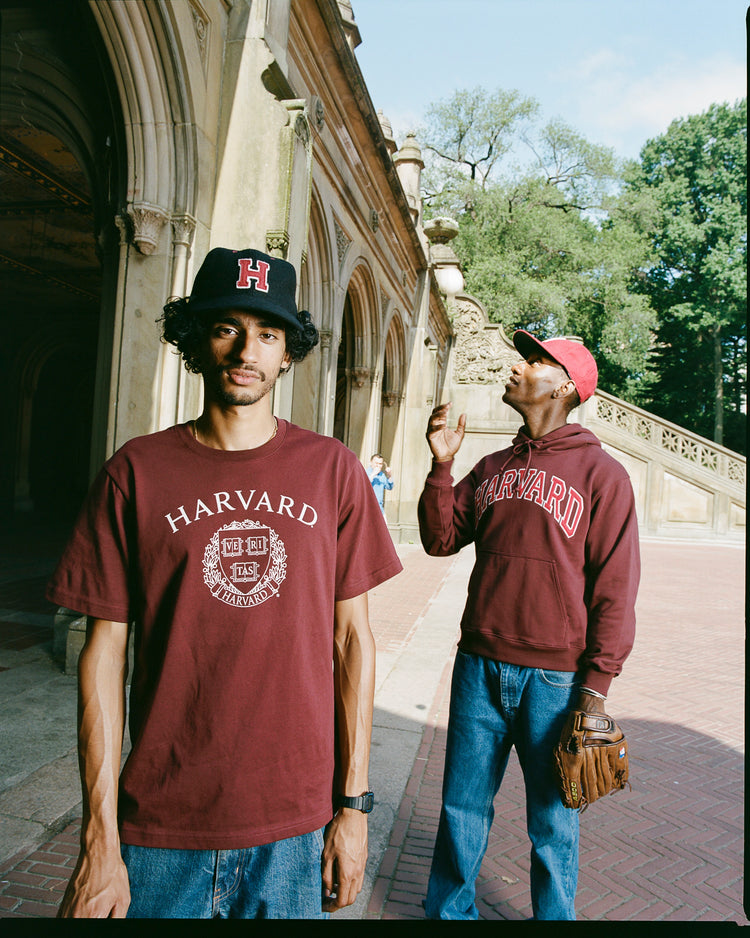 Two men in maroon Harvard attire stand outside an ornate stone building. The man behind wears the EFF Harvard University Crimson H Authentic Wool Ballcap and holds a baseball glove, while lush trees frame the background.