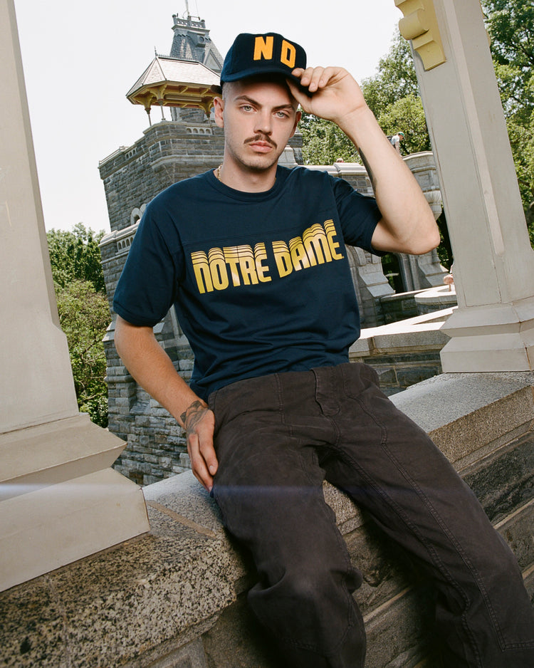 A man sits on a stone ledge outdoors, wearing a navy Notre Dame T-shirt, dark pants, and the EFF University of Notre Dame 1966 Authentic Wool Ballcap, with yellow ND letters. Trees and a stone building are in the background.
