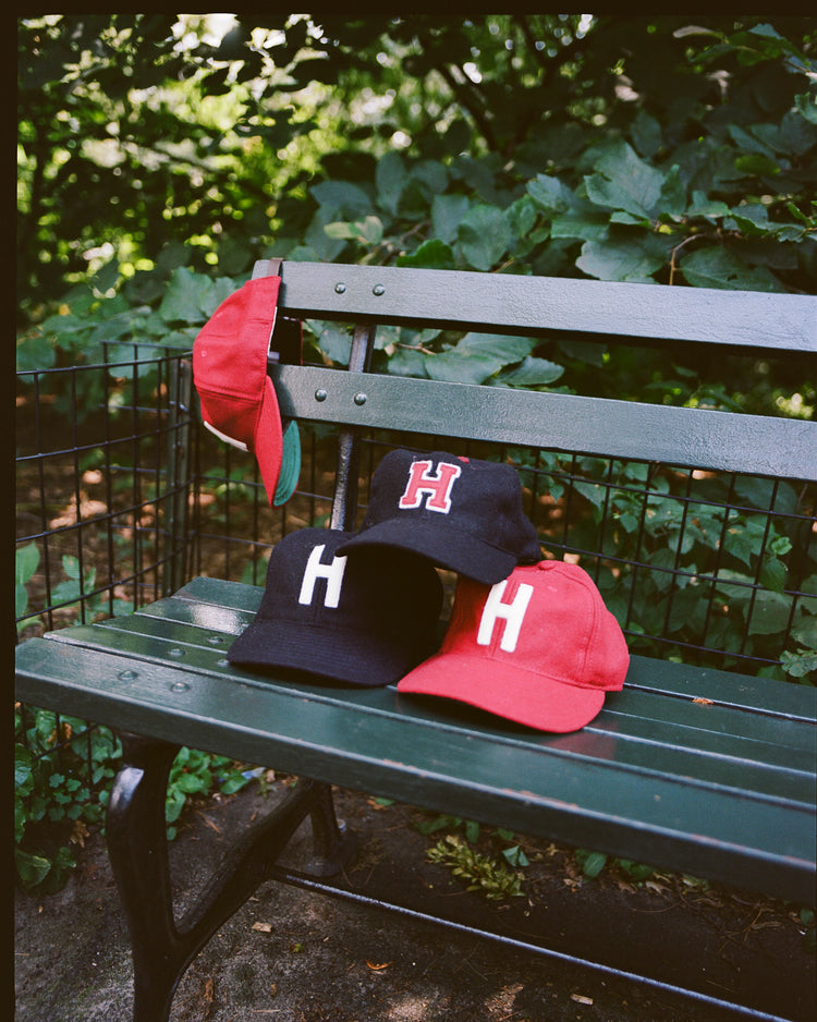 Four vintage EFF Harvard University Crimson H Authentic Wool Ballcaps—two red, two black—each featuring a large H on the front, are displayed on and behind a green park bench amid lush greenery.