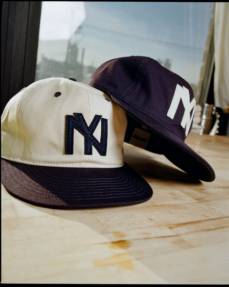 Two Ebbets Field Brooklyn Eagles 1935 Cotton Twill Ballcaps—one white with a dark brim, one navy—display bold NY logos on a light wooden surface, framed by a window and cityscape in the background.