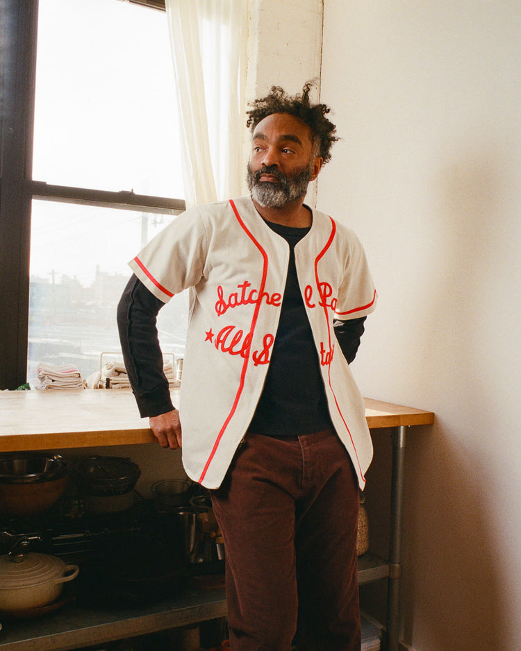 A bearded man with curly hair stands in a sunlit kitchen, wearing an Ebbets Field Satchel Paige All-Stars 1933 Cotton Twill Jersey with red lettering over a black shirt and brown pants, resting his hands on a wooden countertop near the window.