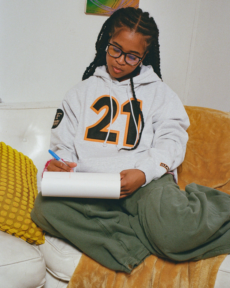 A person with long braids and glasses sits cross-legged on a couch, wearing green pants and the Ebbets Field Karabo Poppy x Ebbets Field Roberto Clemente Heavyweight Cotton Pullover Hoodie, writing in a notebook. A yellow pillow and blanket are nearby.
