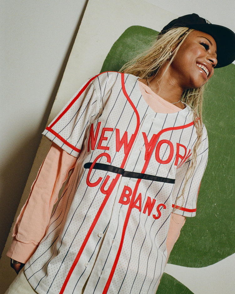 A smiling person wears the Ebbets Field New York Cubans Mesh Baseball Jersey, a long-sleeve shirt, and a black cap while standing before a green and white abstract background.