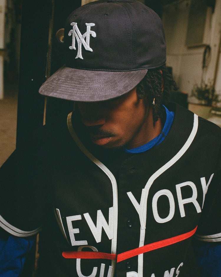 A person wears an Ebbets Field New York Cubans 1948 Cotton Twill Suede Ballcap and a black NY Cubans jersey, honoring historic Negro League teams, while standing indoors with their face partially in shadow.