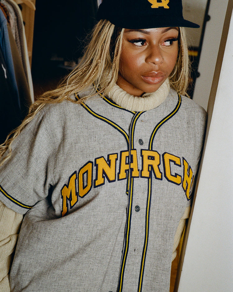 A person with long blonde braids wears the EFF Kansas City Monarchs 1945 Authentic Wool Road Baseball Jersey over a turtleneck and a black cap with a yellow logo, standing indoors next to a rack of clothes.