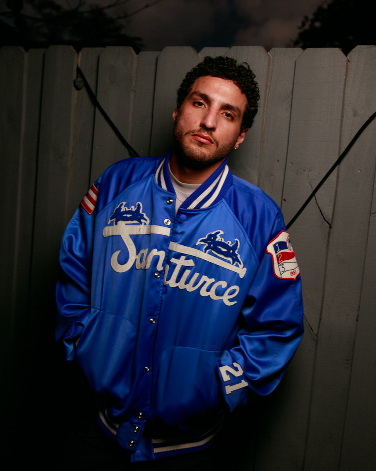 A man with curly hair wears an Ebbets Field Santurce Cangrejeros Cotton Lined Satin Varsity Jacket, standing in front of a wooden fence at night with his hands in his pockets and a neutral expression.