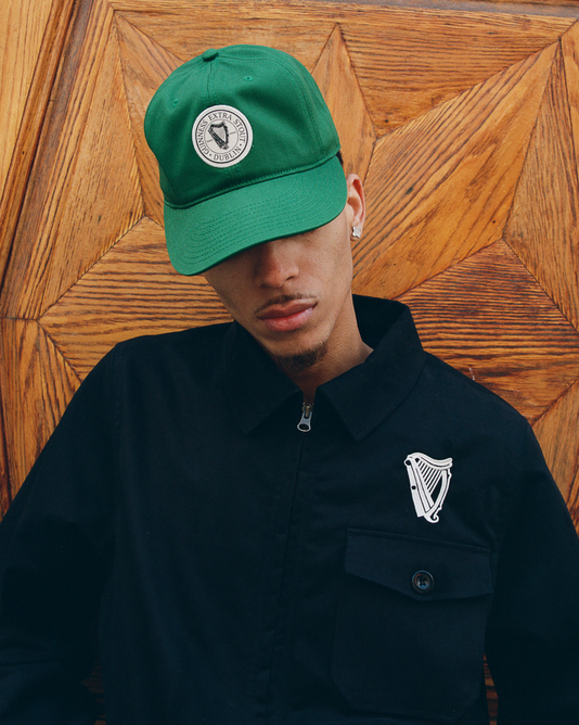 A person wearing the Ebbets Field Guinness Extra Stout Patch Cotton Twill Ballcap, a black jacket with a white harp emblem, and silver hoop earrings stands before a wooden, geometric-patterned background.