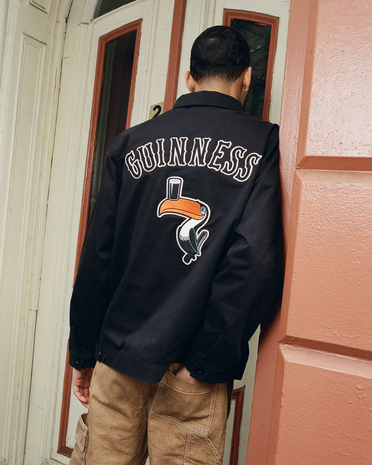 A person in an Ebbets Field Guinness Cotton Grounds Crew Jacket stands with their back to the camera, facing a door. The jacket displays the classic logo and toucan embroidery. A cream door frame and red wall complete the scene.