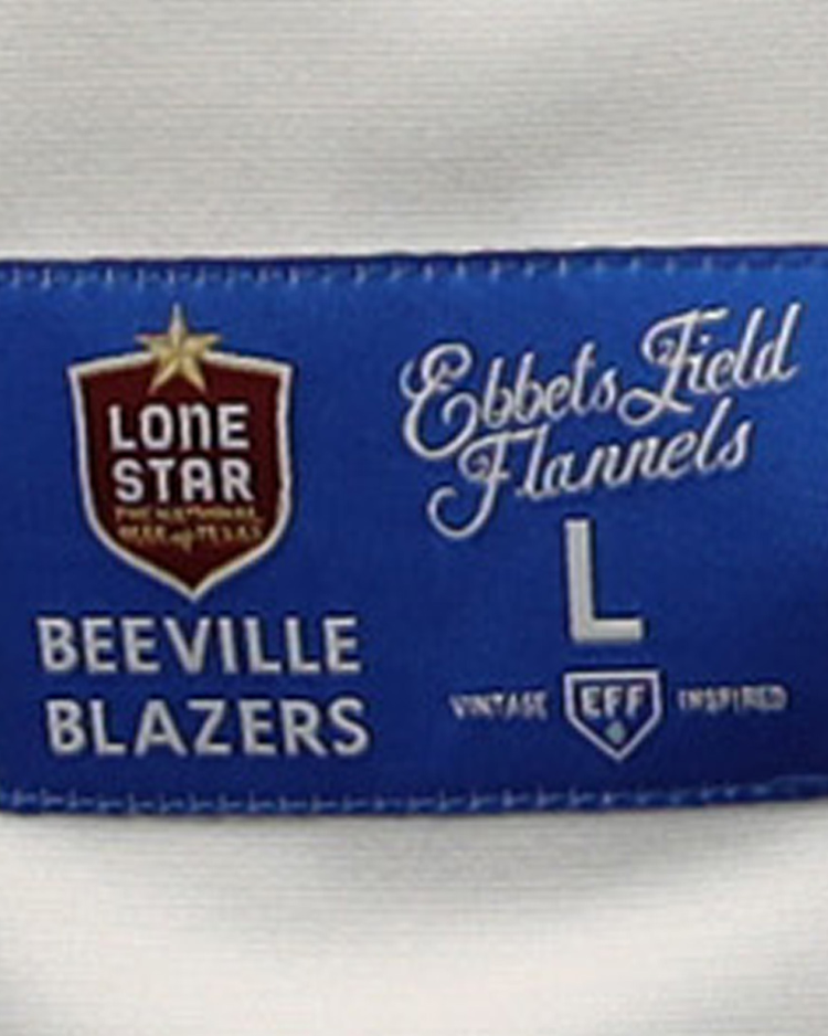The blue and white fabric label on the Beeville Blazers EFF Lone Star Baseball Jersey (by EFF) features various logos, decorative fonts, and text including Beeville Blazers, Joe Hunter Field, L, and Vintage EFF Inspired.