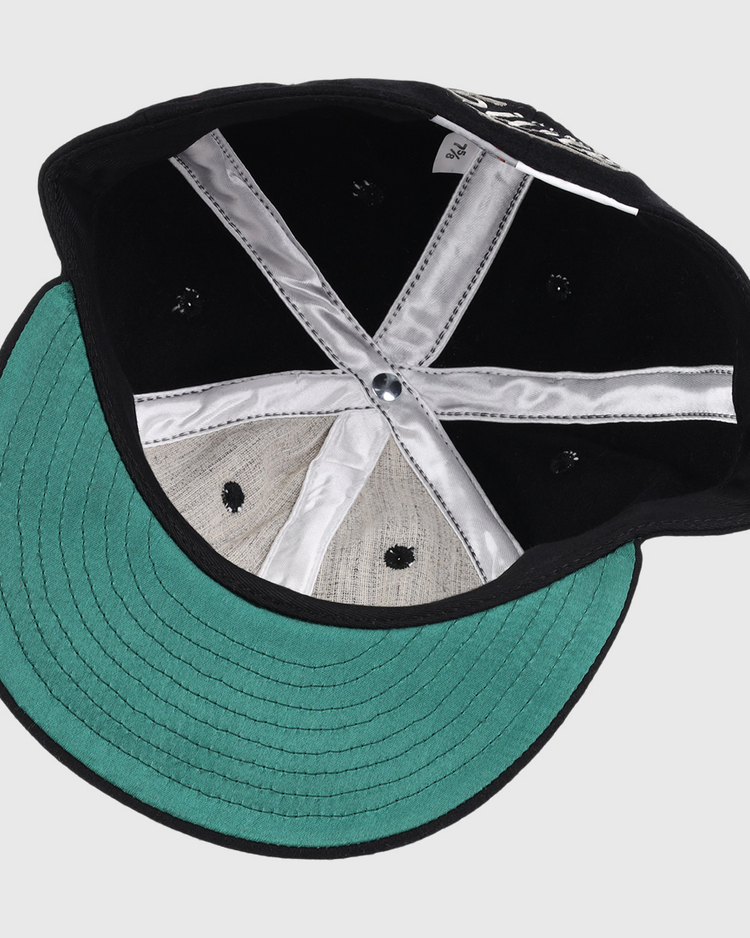 The image shows the inside of an Ebbets Field Flannels Black Fives Wool Ballcap, featuring a green underbrim, silver satin taping, center button, black ventilation holes, and a small white tag near the back.