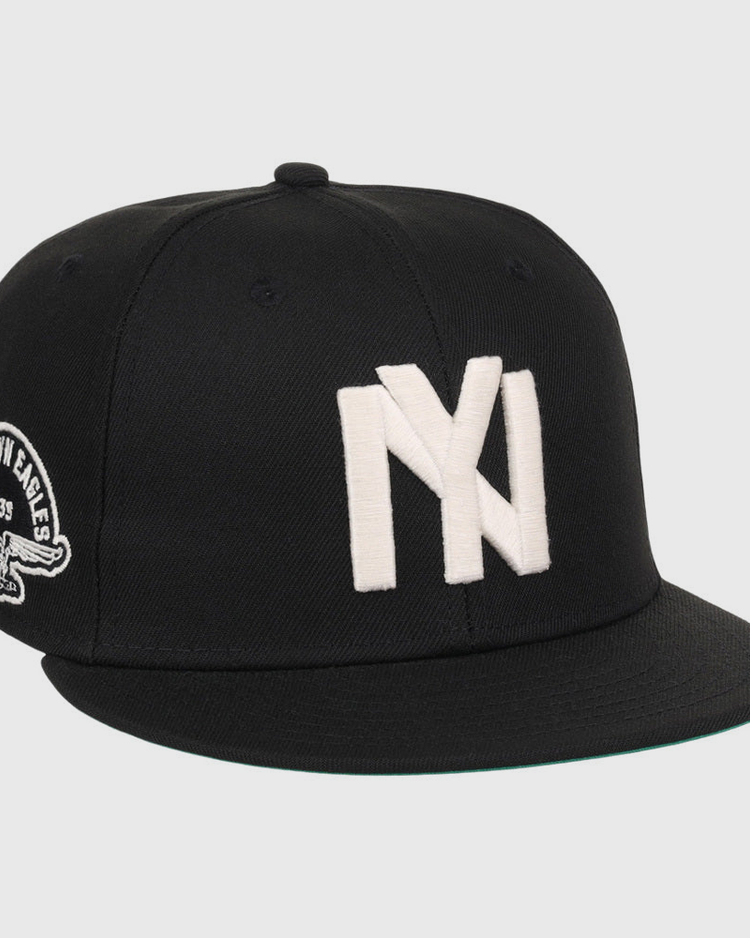 The Ebbets Field Brooklyn Eagles NLB Fitted Ballcap is a black cap with large white overlapping NY letters on the front and a patch showing an eagle and 