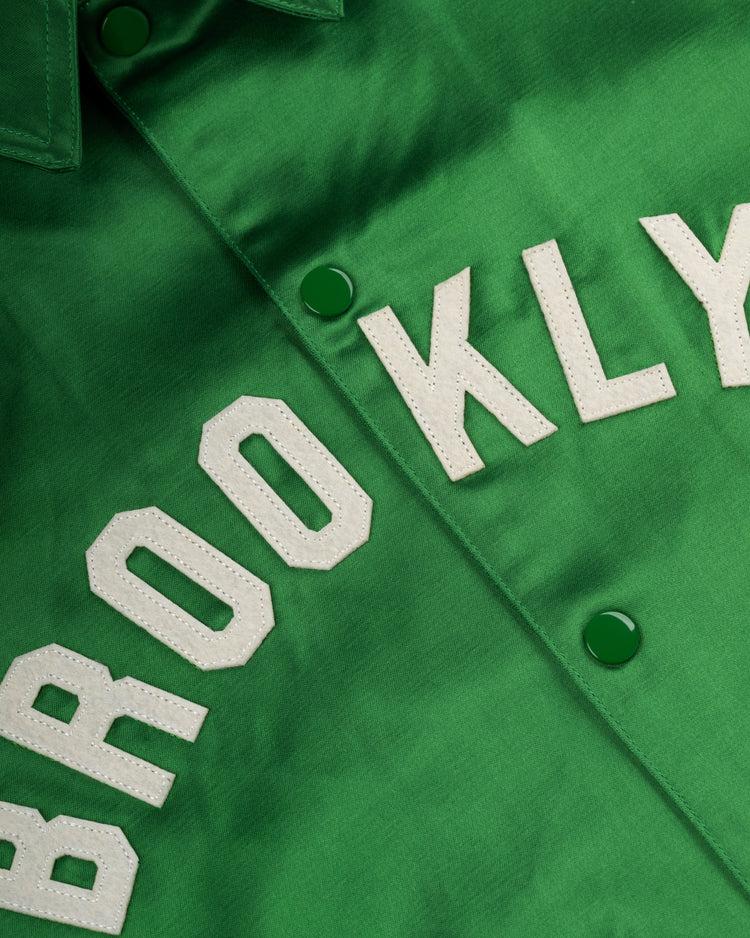 A close-up of the EFF Brooklyn Dodgers 1937 Satin Windbreaker Jacket features green satin, varsity-style white stitched BROOKLYN letters, and classic green snap buttons capturing authentic vintage style.
