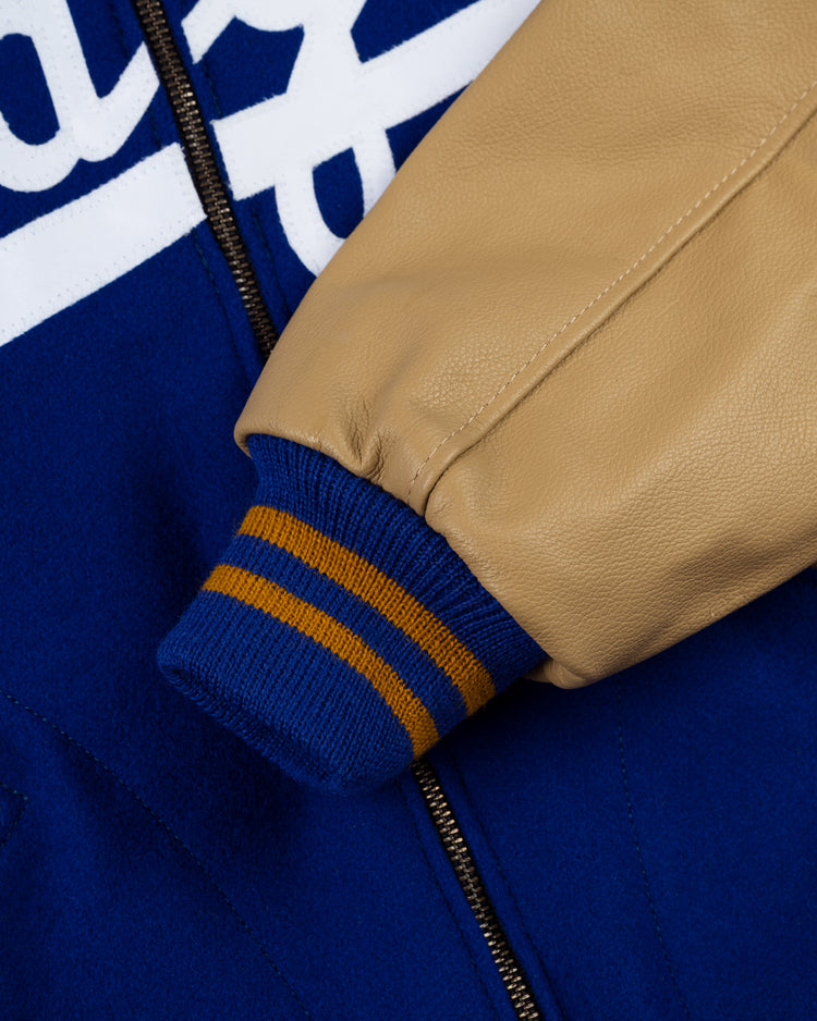 Close-up of the EFF Brooklyn Dodgers 1951 Authentic Wool & Leather Varsity Jacket featuring a beige leather sleeve, blue knit cuff with orange stripes, blue wool body, and part of a white script logo near the zipper.