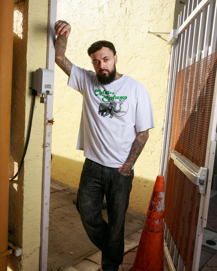 A tattooed man in a Cienfuegos Elefantes Short Sleeve Graphic Tee by Ebbets Field, dark jeans, and white sneakers leans against a yellow wall near a white metal gate and orange traffic cone as sunlight casts shadows.