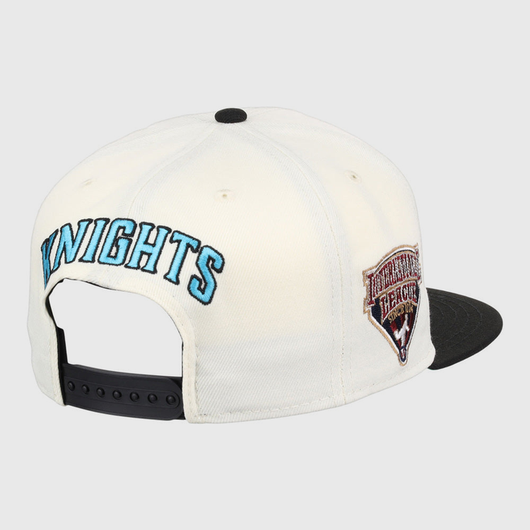 The Ebbets Field Charlotte Knights MiLB Snapback Ballcap features a white design with KNIGHTS embroidered in blue on the back, a HERITAGE HERALD side patch, a black brim, and an adjustable black strap.