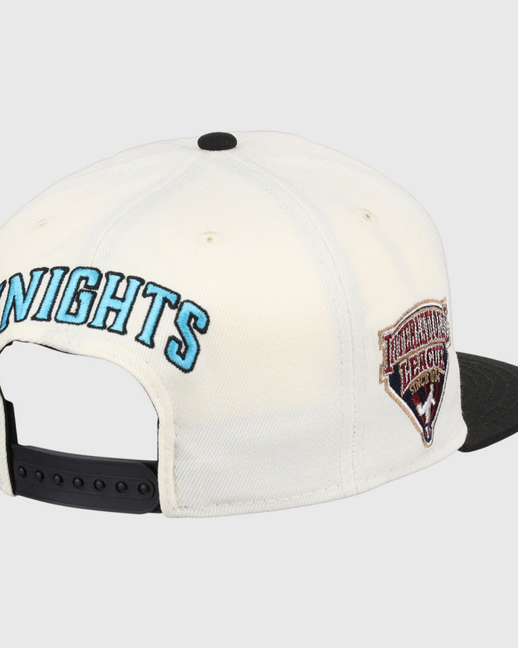 The Ebbets Field Charlotte Knights MiLB Snapback Ballcap features a white design with KNIGHTS embroidered in blue on the back, a HERITAGE HERALD side patch, a black brim, and an adjustable black strap.