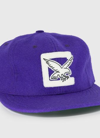 The EFF Cleveland Falcons 1936 Vintage Ballcap is a purple wool cap with a white patch and embroidered flying eagle on the front, a circular black and white logo on the side, and an adjustable leather strap for a custom fit.