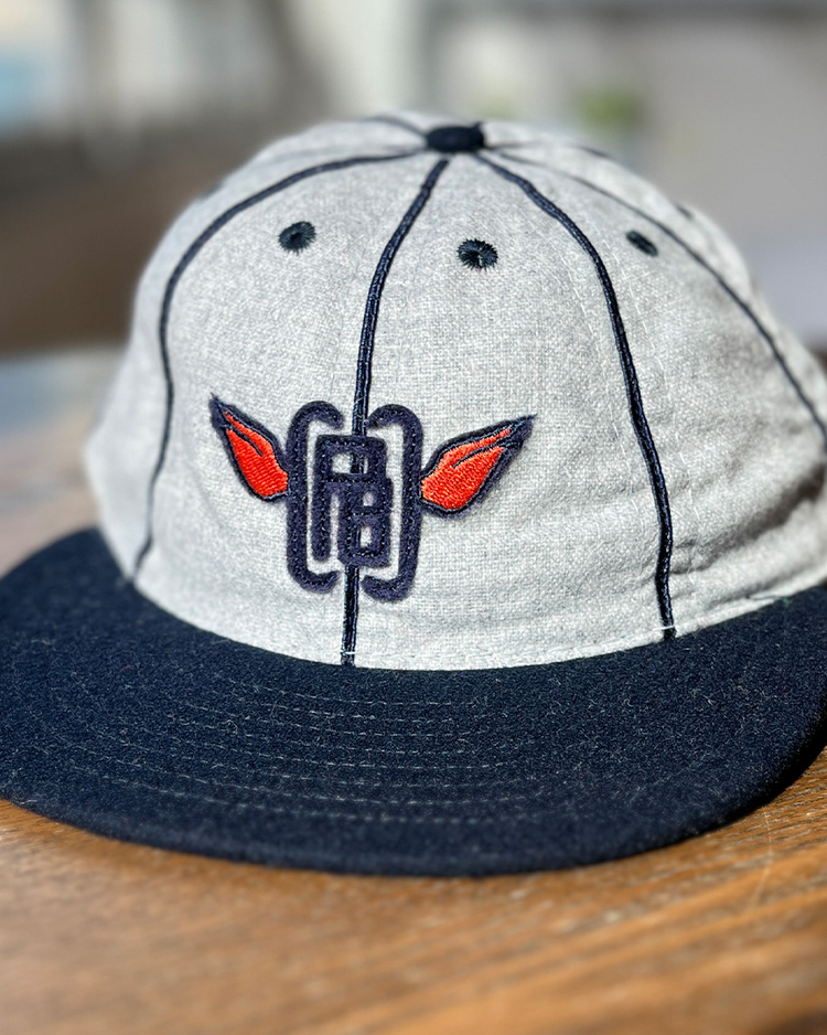 The EFF Columbia Park 1909 Vintage 8-Panel Ballcap in gray and navy, with a dark brim and embroidered logo of red wings and blue lines, sits on a wooden surface.