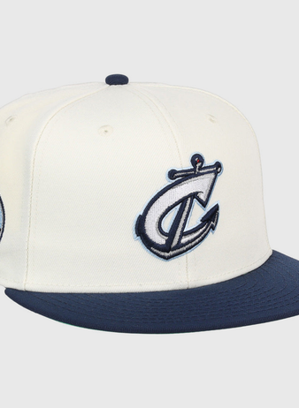 The Ebbets Field Columbus Clippers MiLB Snapback Ballcap is a white hat with a navy blue brim, featuring a stylized anchor and C logo on the front and a circular 614 cityscape patch on the side.