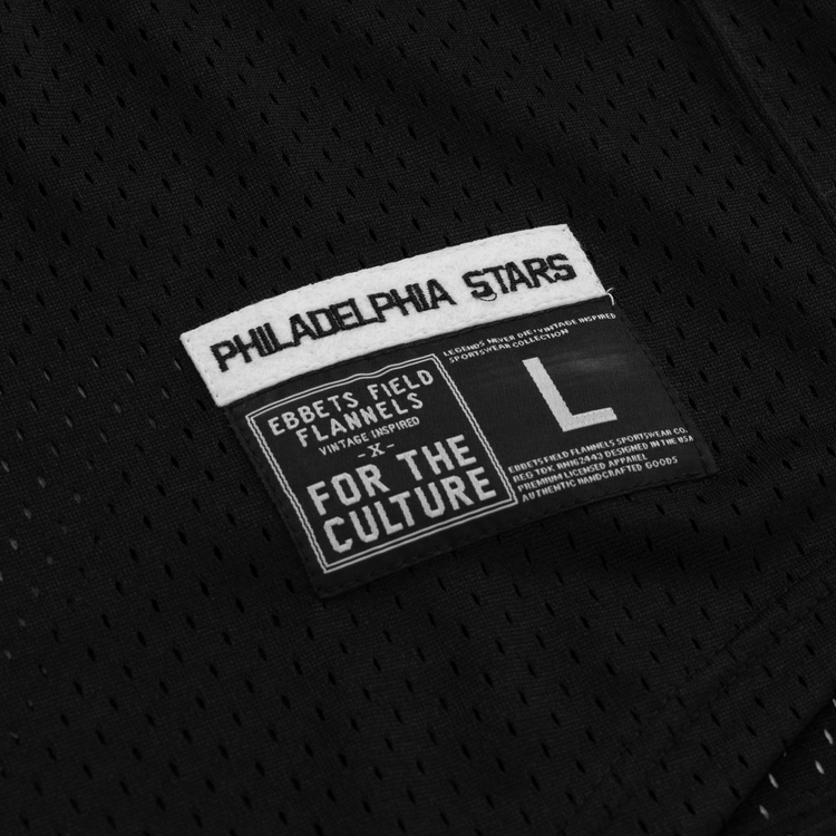 A close-up of black mesh fabric with two labels: one celebrates the Philadelphia Stars and EFF’s Cultural Excellence x Philadelphia Stars Button Down Jersey, while the other highlights Ebbets Field Flannels, For the Culture, and size L.