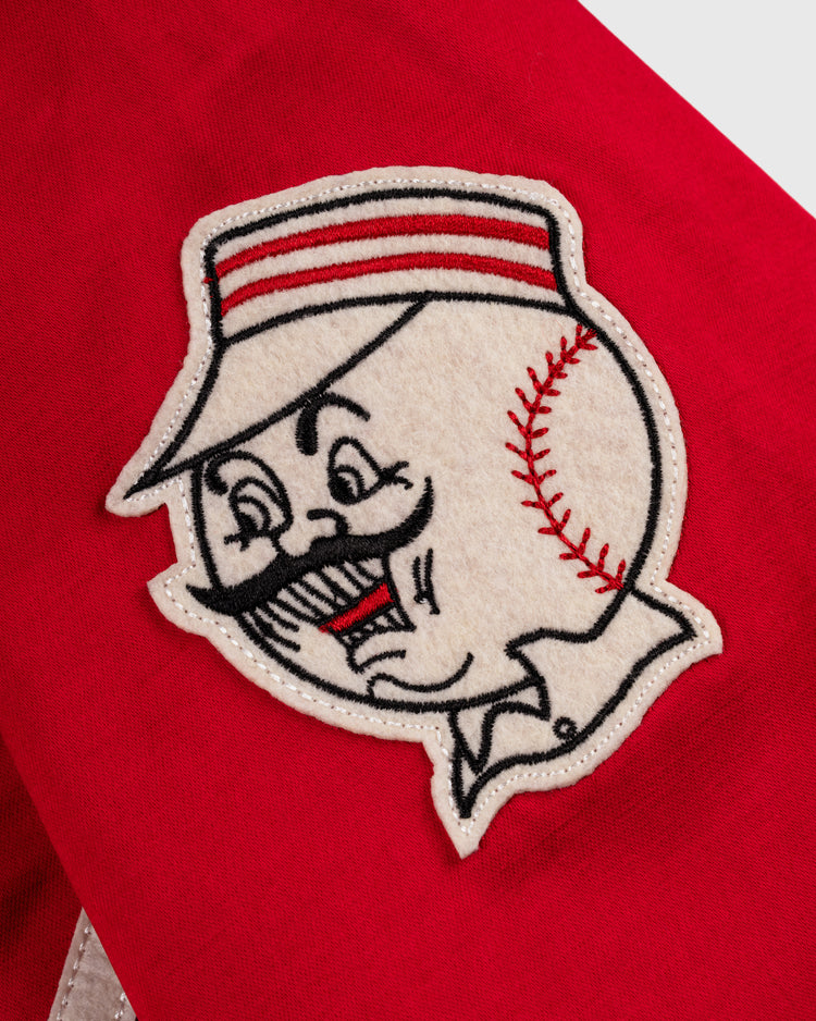 A close-up of the EFF Cincinnati Reds 1955 Satin Windbreaker Jacket shows a red fabric patch featuring a mustached cartoon baseball character with visible stitching and a striped cap.