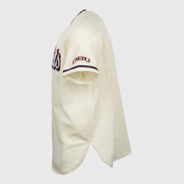 Side view of the EFF Dallas Baptist University Home Jersey: short-sleeve, cream-colored with red and blue accents. DBU is embroidered on the sleeve, a script logo appears on the chest, and it features a split bottom hem.