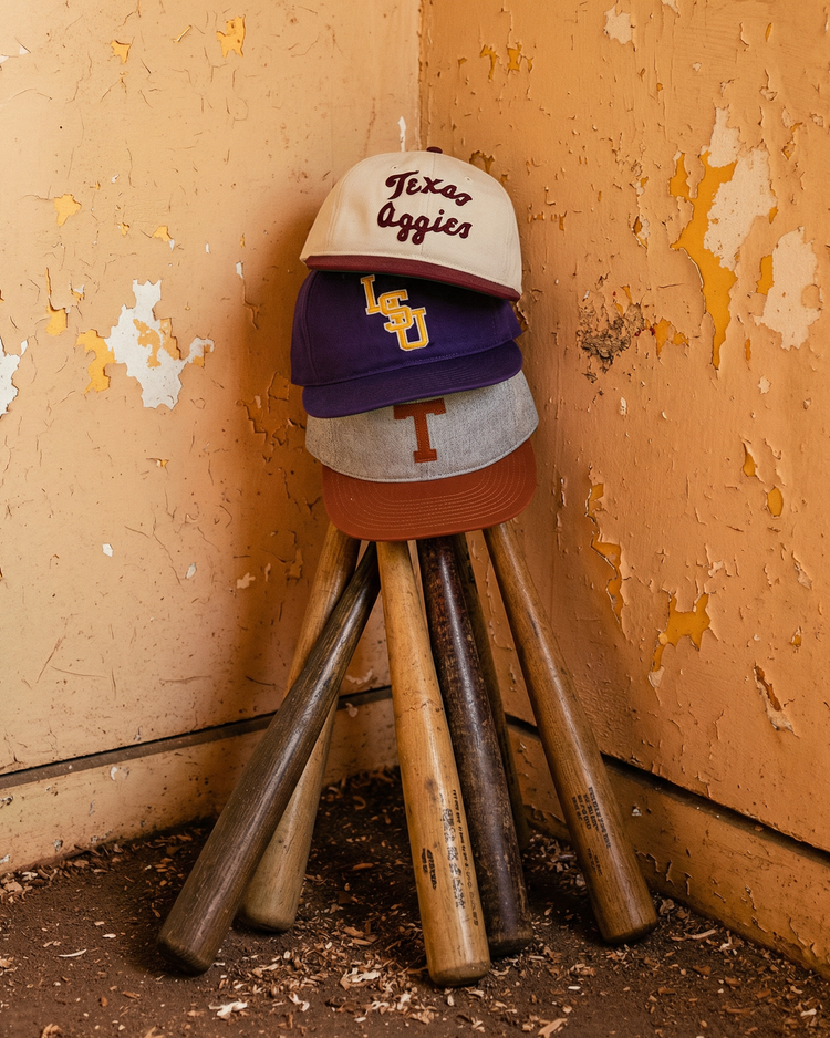 Three baseball bats lean in a corner with three vintage ballcaps stacked on top, including an Ebbets Field Texas A&M University 1952 Cotton Twill Ballcap, plus LSU and burnt orange styles. The paint on the walls is chipped and peeling.