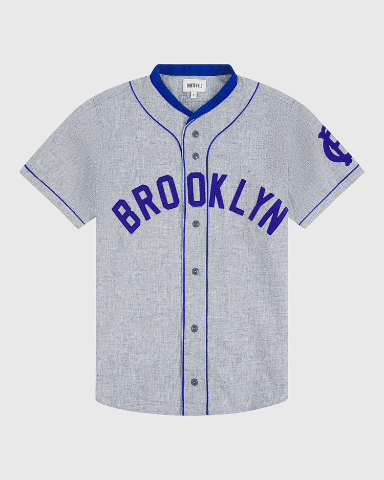 The Ebbets Field Brooklyn Royal Giants 1915 Authentic Wool Road Jersey features a gray short-sleeve design with blue piping and BROOKLYN in bold blue letters across the chest, inspired by the classic 1915 team jersey. It has a blue logo on the left sleeve and front button closure.