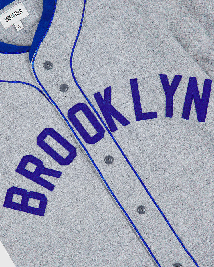 The Ebbets Field Brooklyn Royal Giants 1915 Authentic Wool Road Jersey features a classic gray wool design with blue trim and purple lettering that spells “BROOKLYN” diagonally across the front. Four central buttons complete the iconic look inspired by the original 1915 uniform.