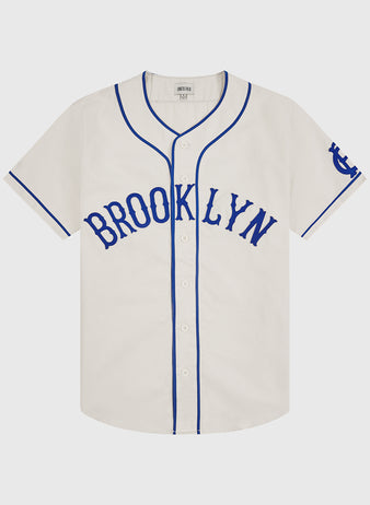 Introducing the Ebbets Field Brooklyn Royal Giants 1919 Cotton Twill Baseball Jersey: a white short-sleeve jersey featuring blue piping, “BROOKLYN” in blue across the chest, blue trim on the sleeves, and a blue logo on the left sleeve—faithfully inspired by the classic 1919 Brooklyn Royal Giants baseball uniform.