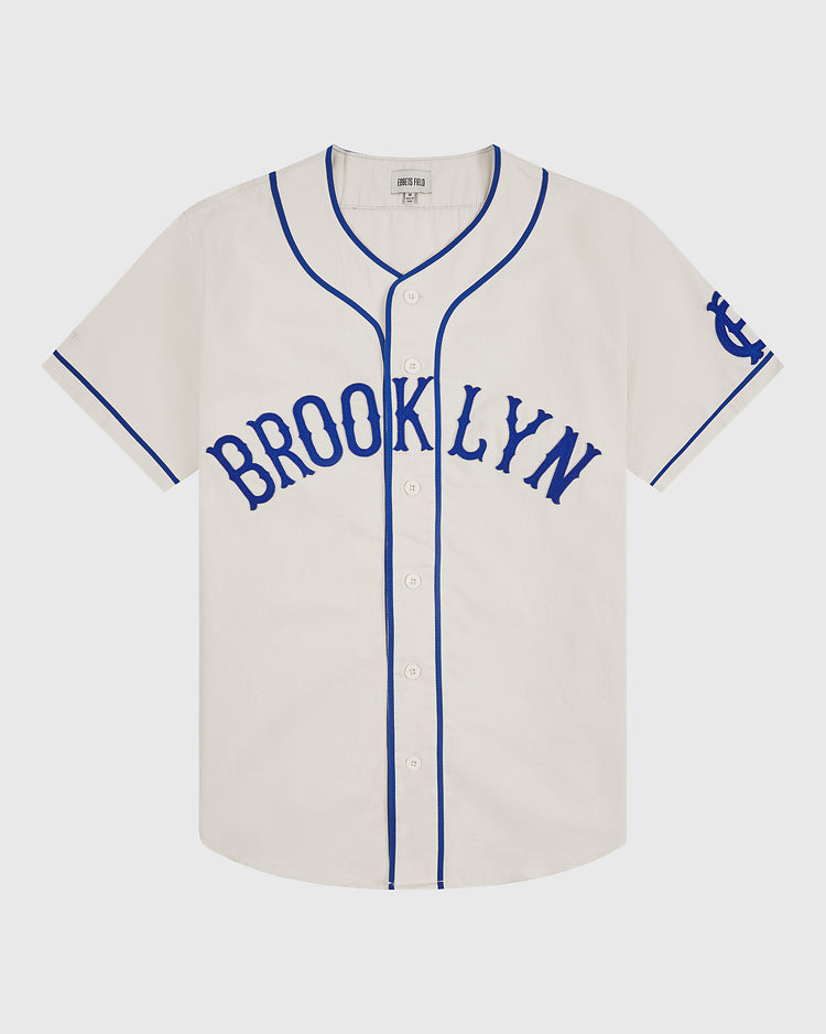 Introducing the Ebbets Field Brooklyn Royal Giants 1919 Cotton Twill Baseball Jersey: a white short-sleeve jersey featuring blue piping, “BROOKLYN” in blue across the chest, blue trim on the sleeves, and a blue logo on the left sleeve—faithfully inspired by the classic 1919 Brooklyn Royal Giants baseball uniform.