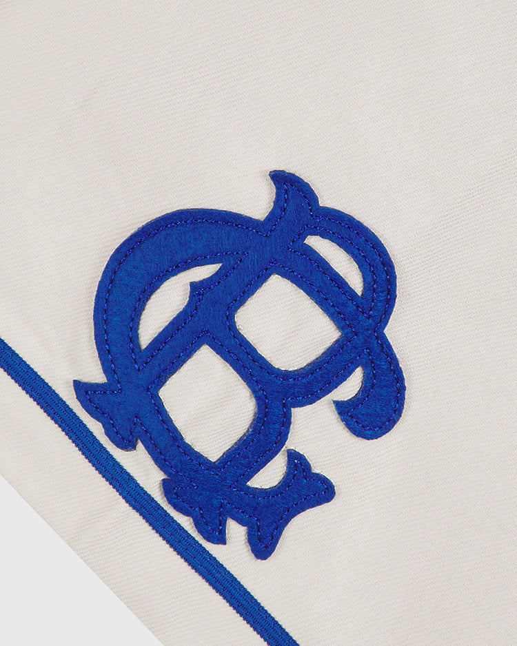 A close-up of the Ebbets Field Brooklyn Royal Giants 1919 Cotton Twill Baseball Jersey showcases a white cotton twill fabric with a blue embroidered letter B, outlined in dark blue and adorned with decorative curls and a blue stripe along the edge, capturing the classic style of the original 1919 uniform.