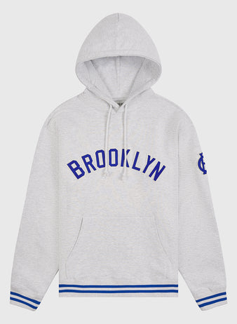 Ebbets Field’s Brooklyn Royal Giants Heavyweight Cotton Terry Pullover Hoodie is crafted from light gray heavyweight cotton, featuring bold blue “BROOKLYN” lettering across the chest, blue and white striped cuffs and hem, a practical front pocket, and a blue emblem on the left sleeve—a tribute to Black baseball history inspired by the legendary Brooklyn Royal Giants.