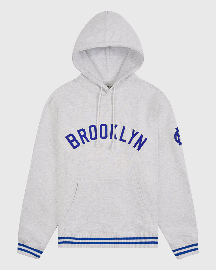 Ebbets Field’s Brooklyn Royal Giants Heavyweight Cotton Terry Pullover Hoodie is crafted from light gray heavyweight cotton, featuring bold blue “BROOKLYN” lettering across the chest, blue and white striped cuffs and hem, a practical front pocket, and a blue emblem on the left sleeve—a tribute to Black baseball history inspired by the legendary Brooklyn Royal Giants.