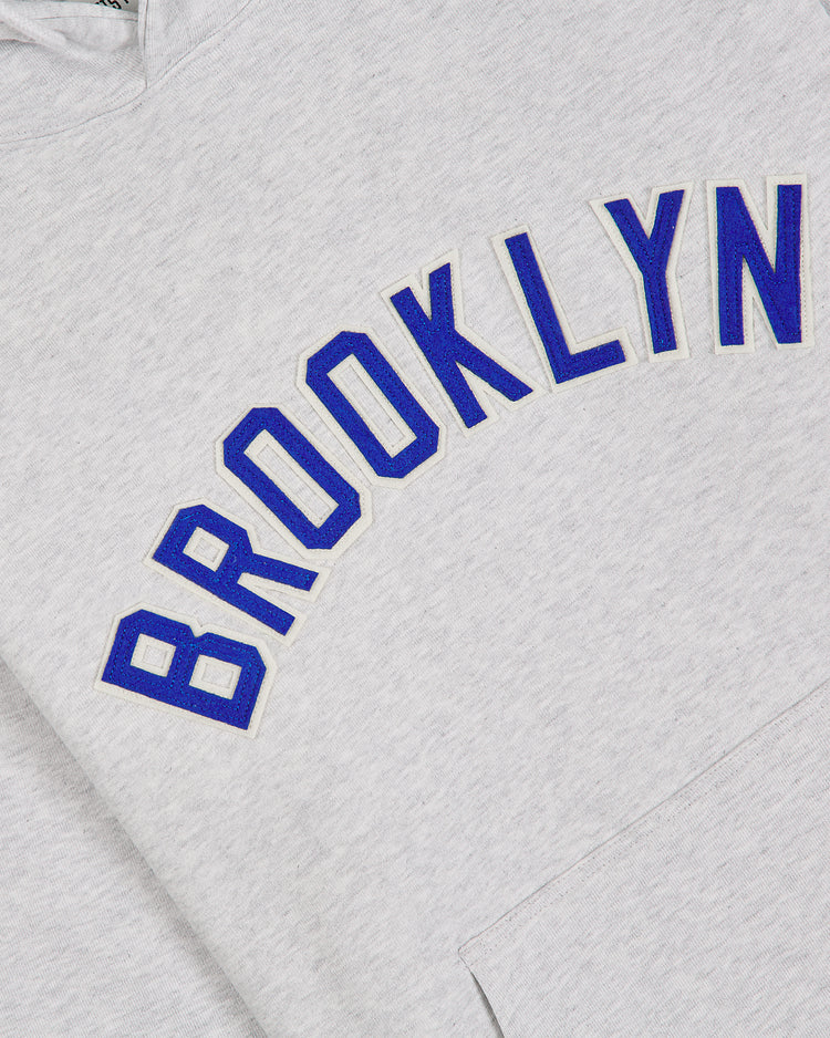 The Ebbets Field Brooklyn Royal Giants Heavyweight Cotton Terry Pullover Hoodie is crafted from heavyweight cotton in grey and features “BROOKLYN” in bold blue letters outlined in white, arched across the chest—a design inspired by Black baseball history and the legacy of the Brooklyn Royal Giants.