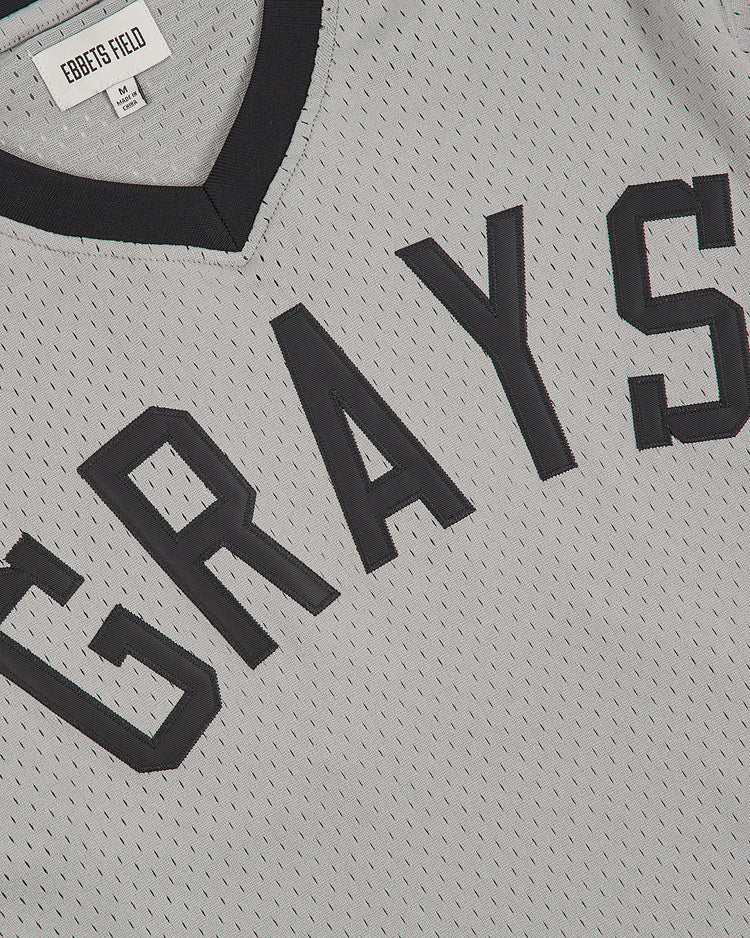 A gray Homestead Grays V-Neck Mesh Jersey by Ebbets Field, featuring black trim and bold black “GRAYS” lettering across the chest, reminiscent of a classic Negro Leagues style. Inside the collar is a white tag marked with “Ebbets Field.”.