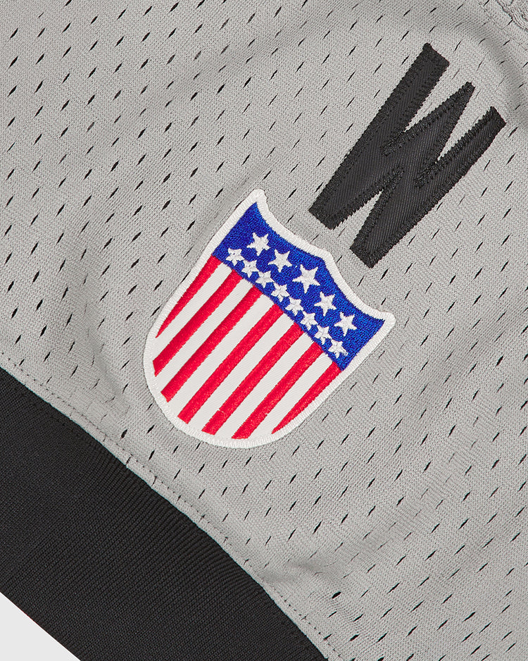 A close-up of the Ebbets Field Homestead Grays V-Neck Mesh Jersey in gray mesh fabric with black trim, showcasing a stitched red, white, and blue shield with stars and stripes and a bold black “W” above it—capturing the timeless style of vintage baseball jerseys inspired by the iconic Negro Leagues’ Homestead Grays.