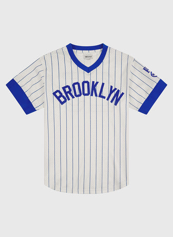 This Brooklyn Royal Giants 1925 V-Neck Mesh Jersey by Ebbets Field features a classic white design with blue pinstripes, complemented by blue trim on the collar and sleeve cuffs. “BROOKLYN” is boldly displayed in blue letters across the chest, while a small commemorative patch adorns the left sleeve. Inspired by timeless Negro Leagues apparel, this vintage-style jersey captures an authentic and enduring look.