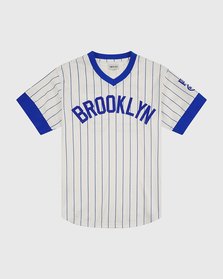 This Brooklyn Royal Giants 1925 V-Neck Mesh Jersey by Ebbets Field features a classic white design with blue pinstripes, complemented by blue trim on the collar and sleeve cuffs. “BROOKLYN” is boldly displayed in blue letters across the chest, while a small commemorative patch adorns the left sleeve. Inspired by timeless Negro Leagues apparel, this vintage-style jersey captures an authentic and enduring look.