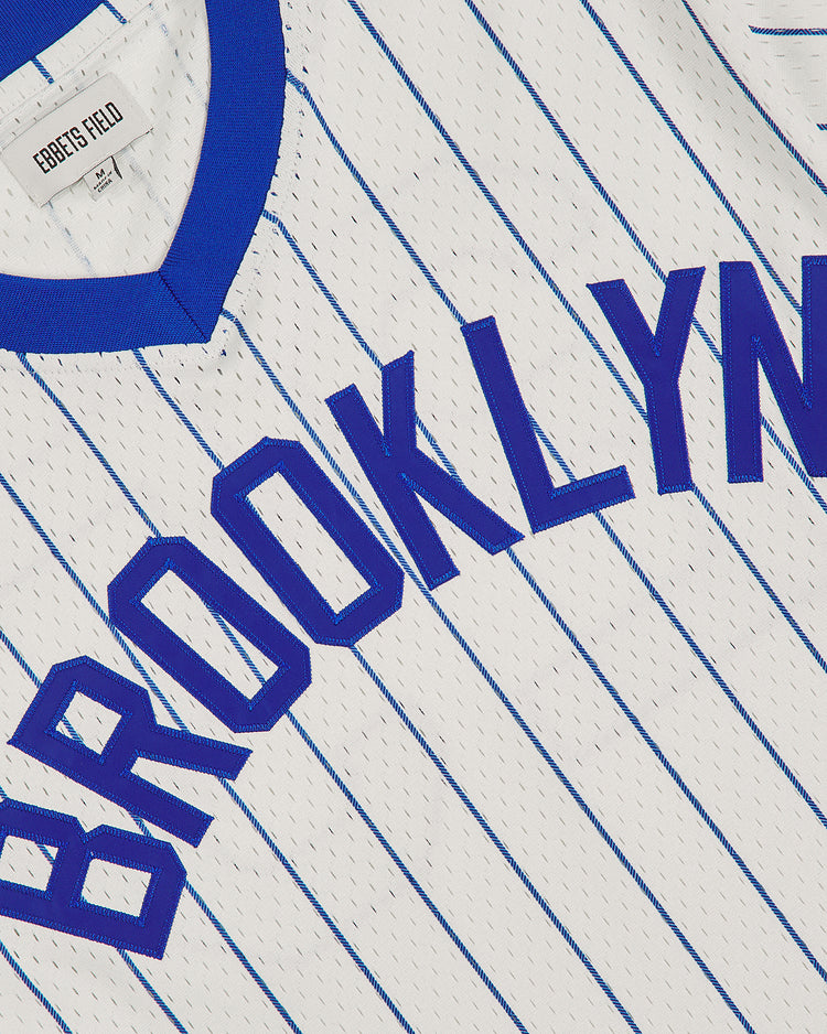 The Brooklyn Royal Giants 1925 V-Neck Mesh Jersey by Ebbets Field features a white design with blue pinstripes, a blue collar, and “BROOKLYN” boldly displayed across the chest. Inspired by classic Negro Leagues apparel, this vintage jersey includes a tag near the collar that reads “Ebbets Field” and “SM.”.