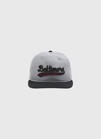 A plain, light gray background showcases the Ebbets Field Baltimore Elite Giants 1949 Cotton Twill Ballcap. This gray cap features a black brim and displays black cursive “Baltimore” text on the front above a bold red strip that reads “Elite Giants,” paying tribute to the historic Baltimore Elite Giants of Negro League baseball.