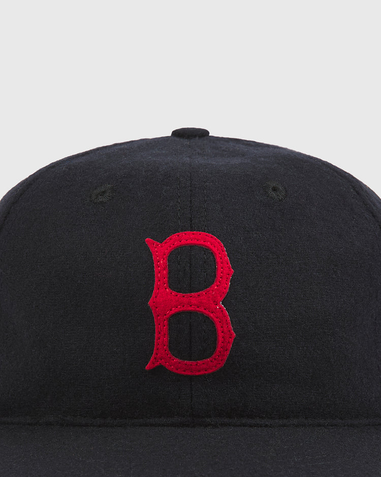 Close-up of the Baltimore Elite Giants 1949 Authentic Wool Ballcap by Ebbets Field, a black baseball cap with a prominent red embroidered letter “B” on the front. The background is plain and light-colored.